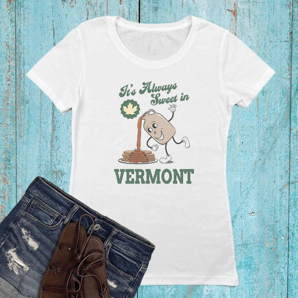 A white short-sleeve graphic T-shirt laid flat on a distressed turquoise wood background, styled with blue jeans and brown lace-up shoes. The shirt graphic reads 'It's Always Sweet in VERMONT' with a cartoon maple syrup pancake and a green maple leaf.