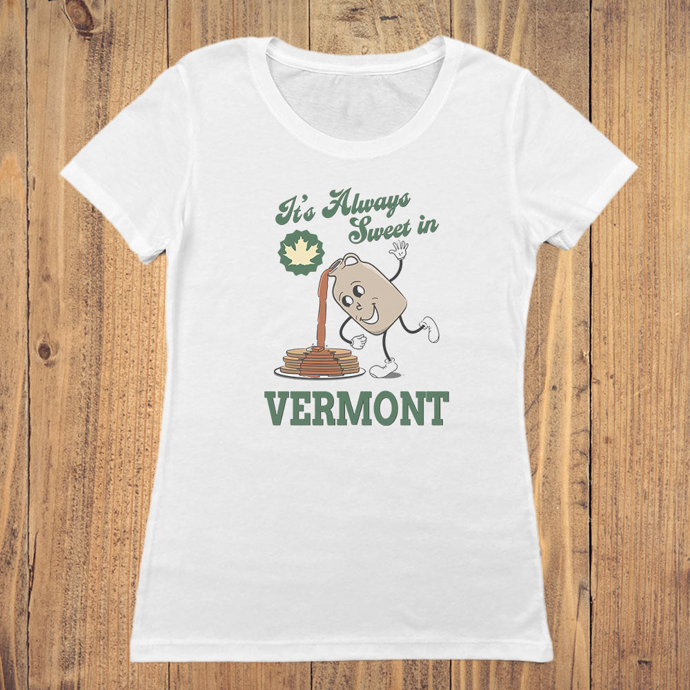 A white short-sleeve crewneck t-shirt with a Vermont-themed graphic print, displayed flat on a rustic wooden plank background. The graphic features the text 'It's Always Sweet in VERMONT' in green, alongside a cartoon maple syrup bottle character and a maple leaf.